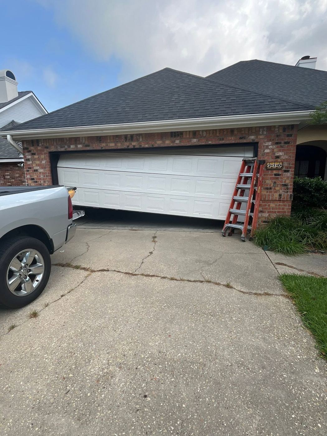 Garage door hanging off track on a brick home, with ladder set up for repair